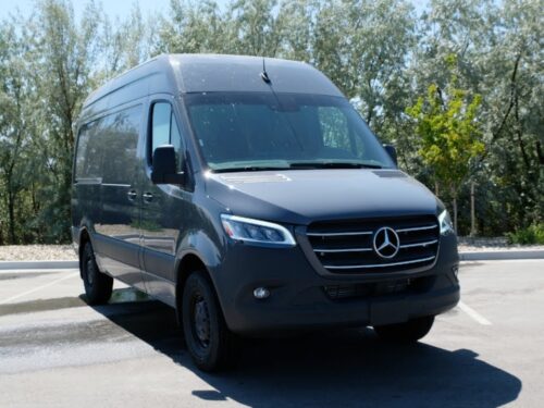 Dark gray Mercedes-Benz Sprinter van parked in a sunny lot with trees in the background